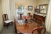 Formal dining room with a polished wooden table and chairs, floral centerpiece, sideboard with mirror, and a curtained window.
