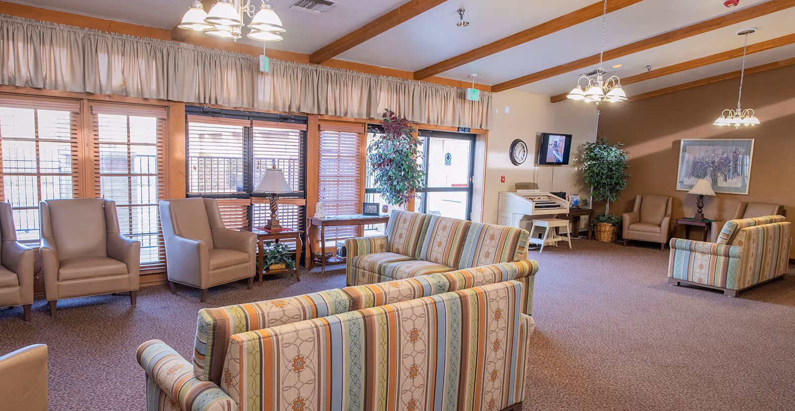 A spacious and well-lit living room area in a retirement community featuring multiple armchairs and striped sofas arranged for conversation. The room has large windows with wooden blinds, beige curtains, carpeted floors, potted plants, and ceiling lights. A clock and a small TV are mounted on the wall, and there is a small table with a lamp and a decorative item.