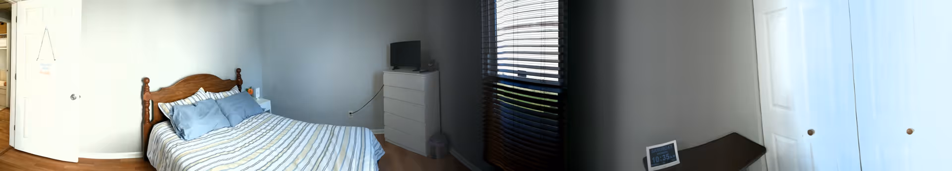 Modest bedroom with a bed dressed in striped bedding, a white dresser topped by a small TV, a window with blinds, and closet doors.