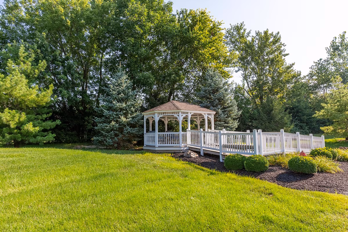A white wooden gazebo with a ramp surrounded by green grass, bushes, and tall trees under a clear sky.