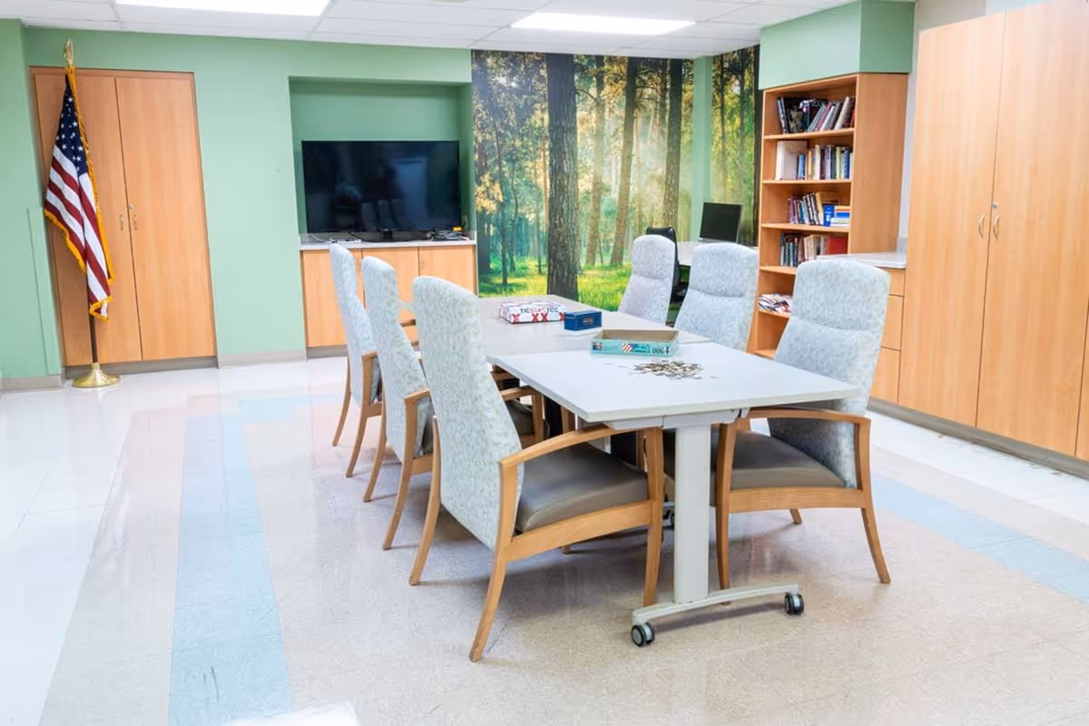 A bright activity room with a central table surrounded by upholstered chairs, a TV, bookshelves, and a forest mural on the back wall.