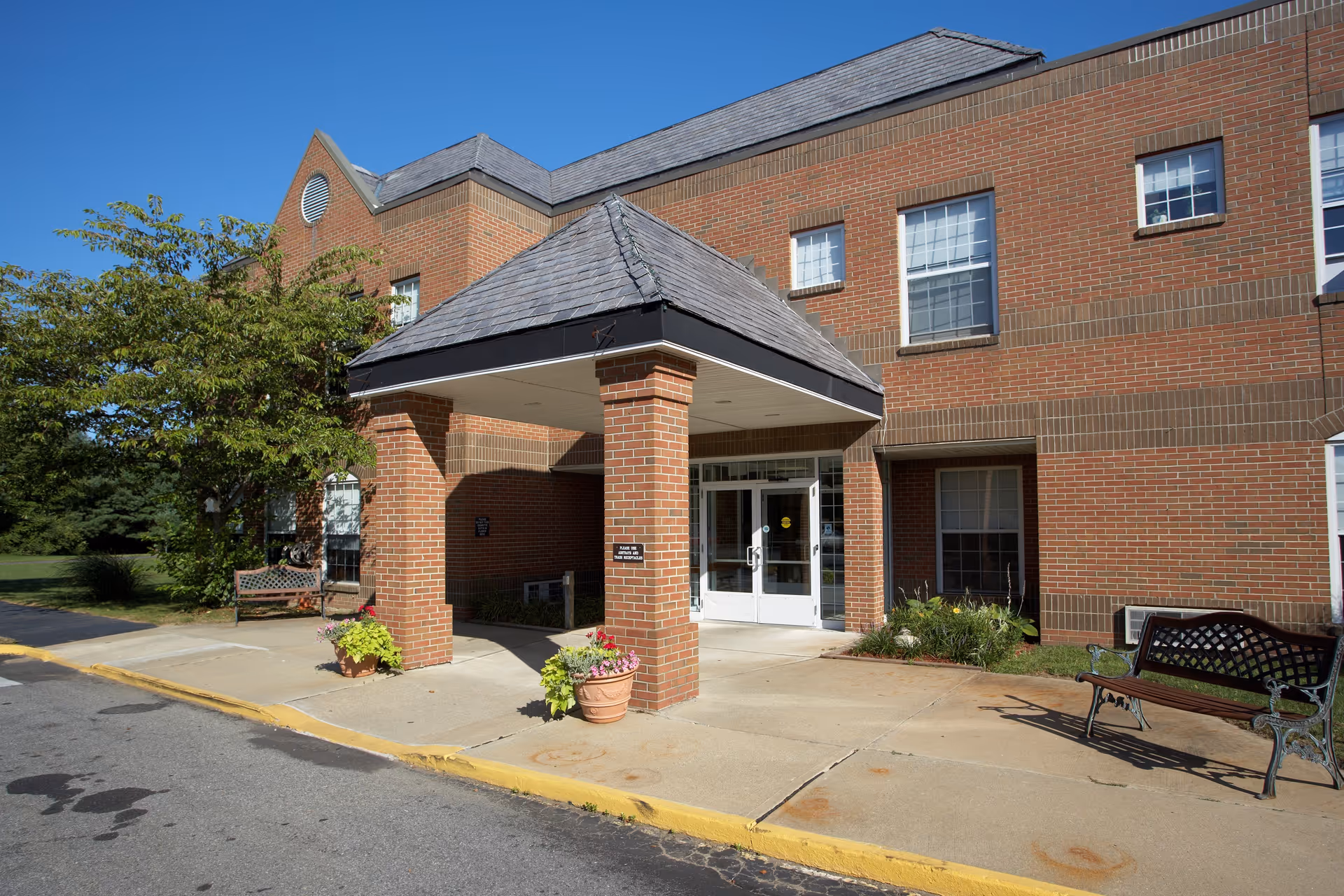 Entrance of a brick building with a covered porch supported by brick columns, two benches on either side, potted plants, and a tree to the left under a clear blue sky.