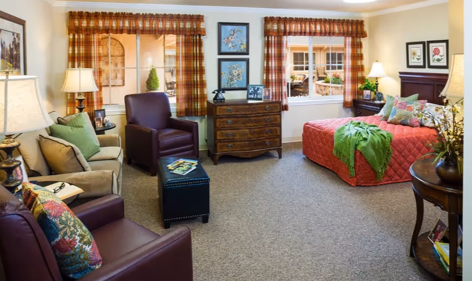 A cozy, well-lit room with a bed covered in a red quilt and green throw, two leather armchairs, a beige sofa with green pillows, a wooden dresser, and windows with plaid curtains. The room is decorated with framed floral artwork, table lamps, and small plants, creating a warm and inviting atmosphere.
