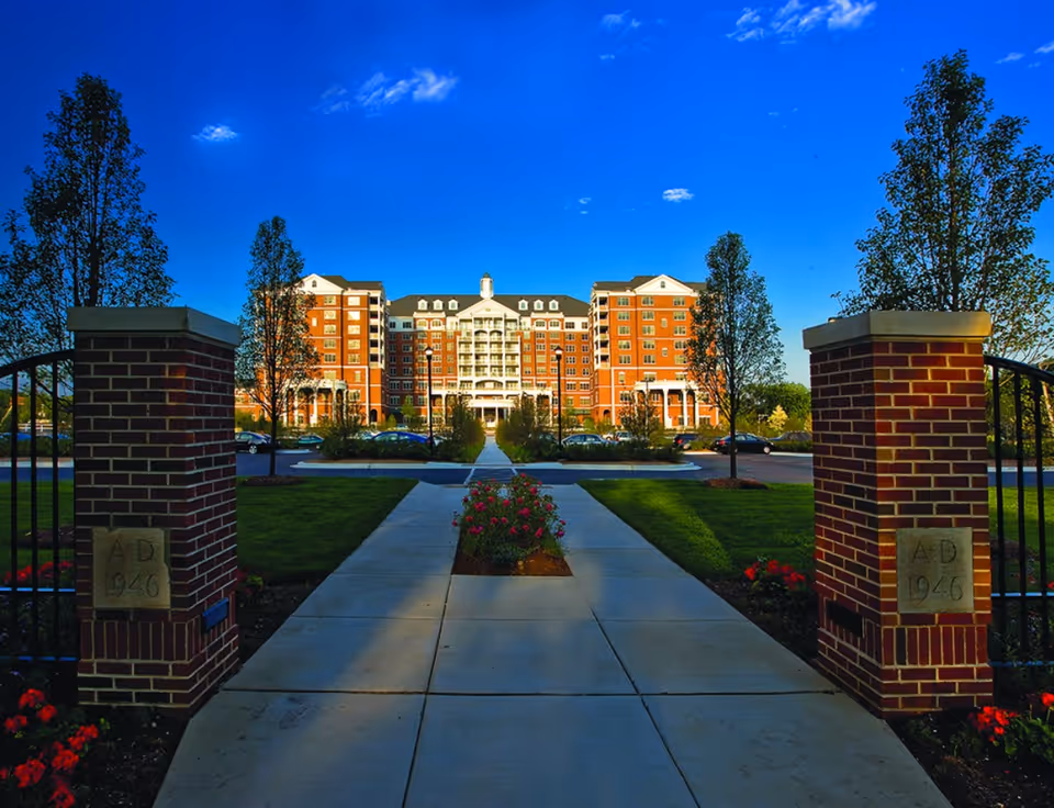 A wide concrete pathway flanked by two brick pillars with plaques reading 'A.D 1946' leads to a large multi-story red brick building with white balconies and a central cupola. The pathway is bordered by green lawns, trees, and flower beds under a bright blue sky with a few clouds.
