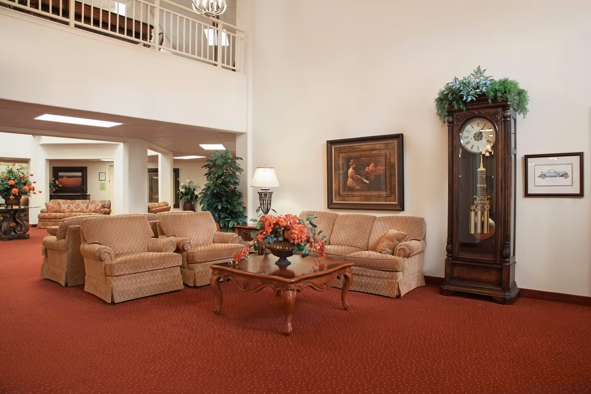 A spacious living room area with beige upholstered sofas and armchairs arranged around a wooden coffee table adorned with a floral centerpiece. The room features a tall grandfather clock decorated with greenery, framed artwork on the walls, a floor lamp, and red carpet flooring. There is a balcony railing visible above and additional seating areas in the background.