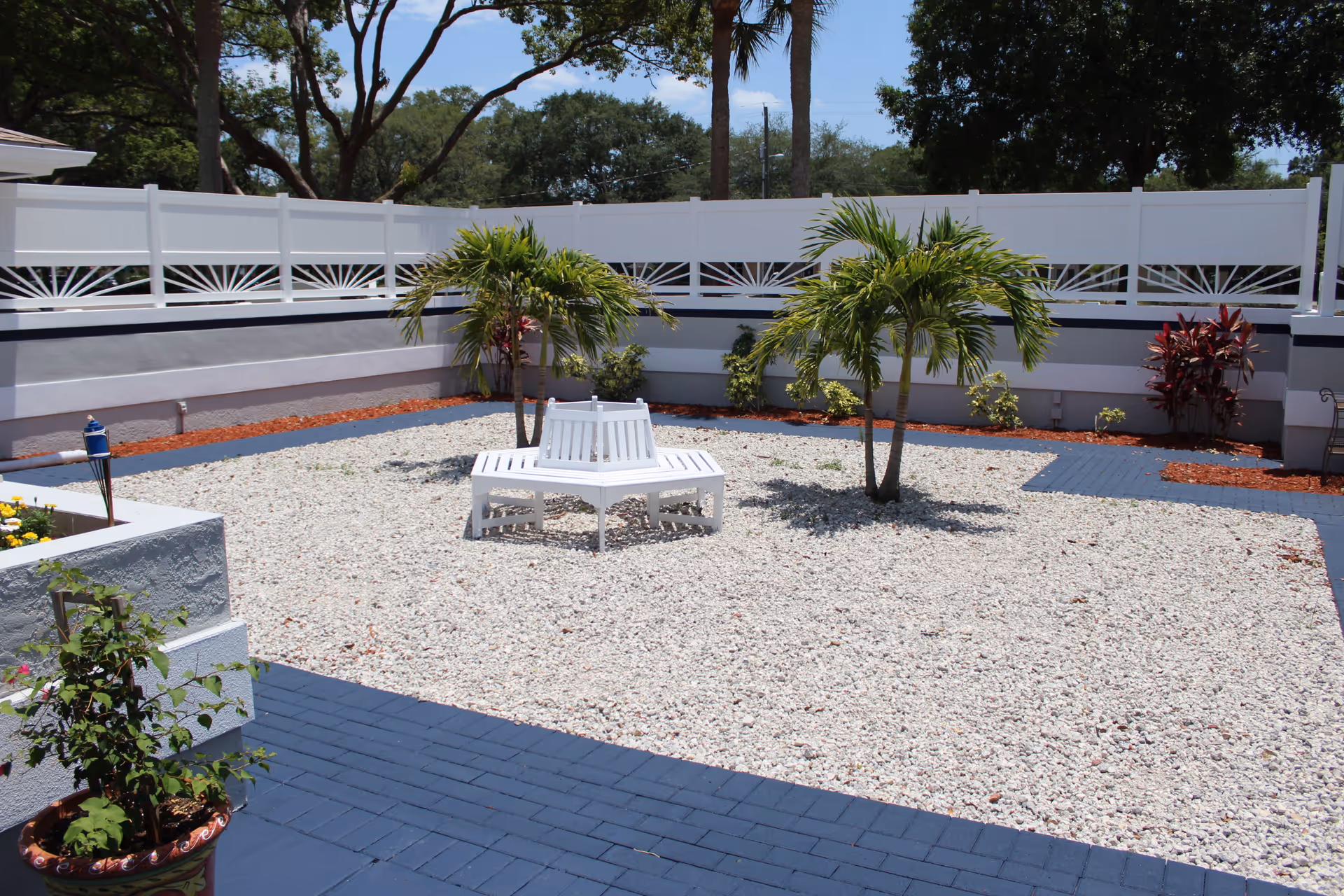 Outdoor courtyard area with white gravel ground cover, two small palm trees, a white circular bench surrounding one of the trees, and a white fence enclosing the space. There are some plants and shrubs along the fence and a potted plant in the foreground.