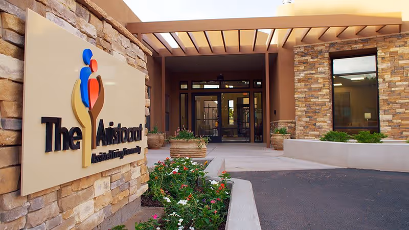 Entrance to The Aristocrat Assisted Living Center with a stone wall sign displaying the facility's name and logo. The entrance features a covered walkway with large glass doors and windows, surrounded by stone and stucco exterior walls. There are flower beds with blooming plants near the entrance.