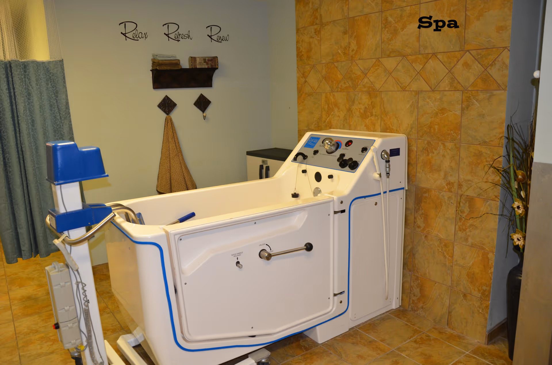 A spa room featuring a white walk-in bathtub with control knobs and a handheld shower head, set against a tiled wall. The wall has the word 'Spa' written on it, and there is a shelf with folded towels and hooks on the adjacent wall with the words 'Relax Refresh Renew' above it. A blue curtain is partially visible on the left side.