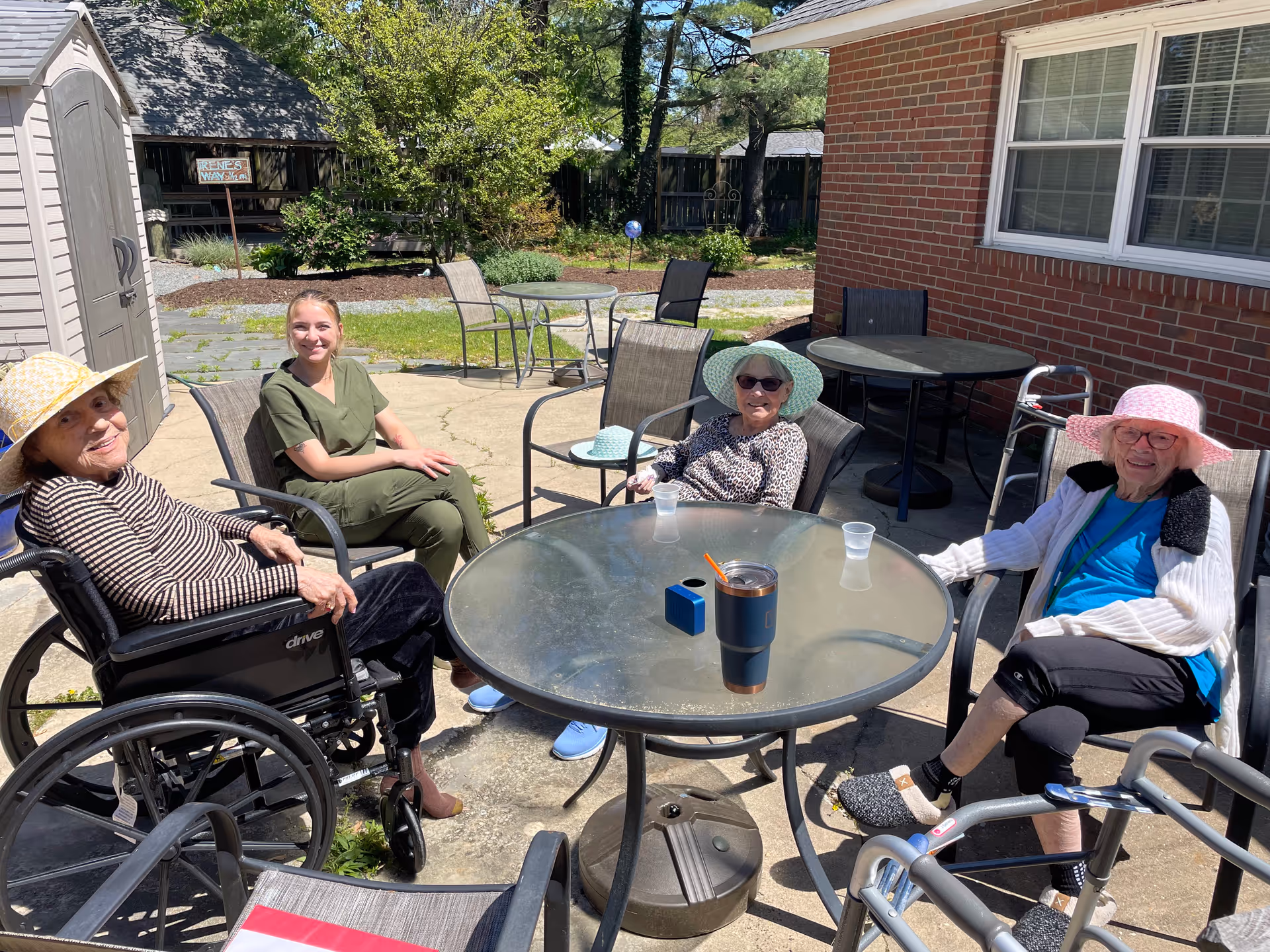 Three elderly women and a caregiver sitting around a round glass outdoor table on a sunny patio. Two women are wearing sun hats and one is in a wheelchair. The caregiver is dressed in green scrubs and smiling. There are drinks on the table and a brick building in the background.