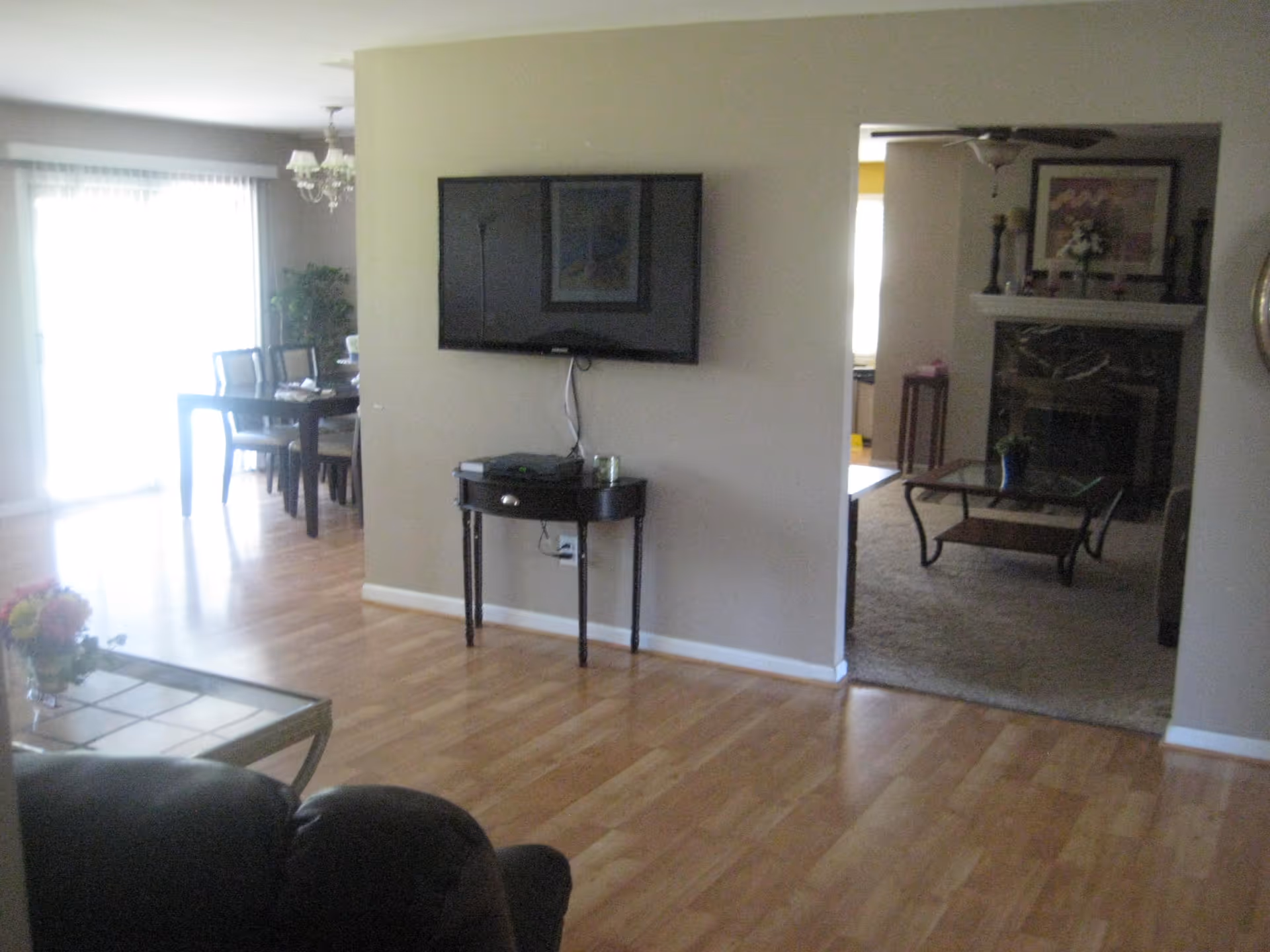 Interior view of a senior living facility showing a living room with a mounted flat-screen TV above a small table, a dining area with a table and chairs near a large window with sheer curtains, and a separate sitting area with a fireplace, coffee table, and framed artwork on the wall.
