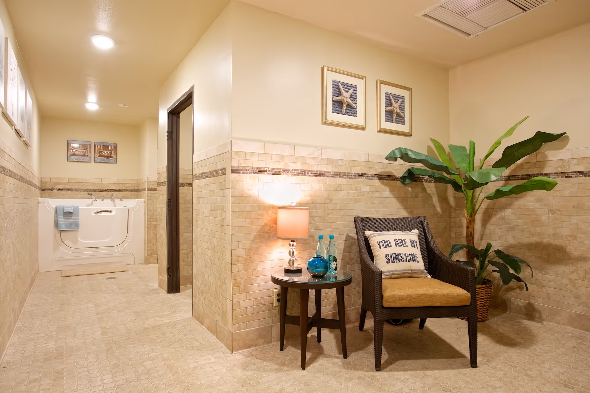A well-lit room with beige tiled walls and floor featuring a walk-in bathtub at the end of the hallway. On the right side, there is a wicker chair with a cushion and a pillow that says 'YOU ARE MY SUNSHINE', next to a small round table with a lamp, two bottles of water, and a decorative blue vase. A large green potted plant is placed beside the chair. Two framed starfish pictures hang on the wall above the chair.