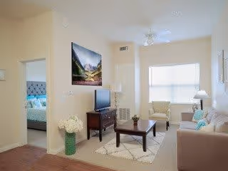 A bright living room with a beige sofa, two armchairs near a large window, a wooden coffee table on a patterned rug, a TV on a wooden stand, a floor lamp, and a vase with white flowers. A bedroom with a bed and blue bedding is visible through an open doorway.