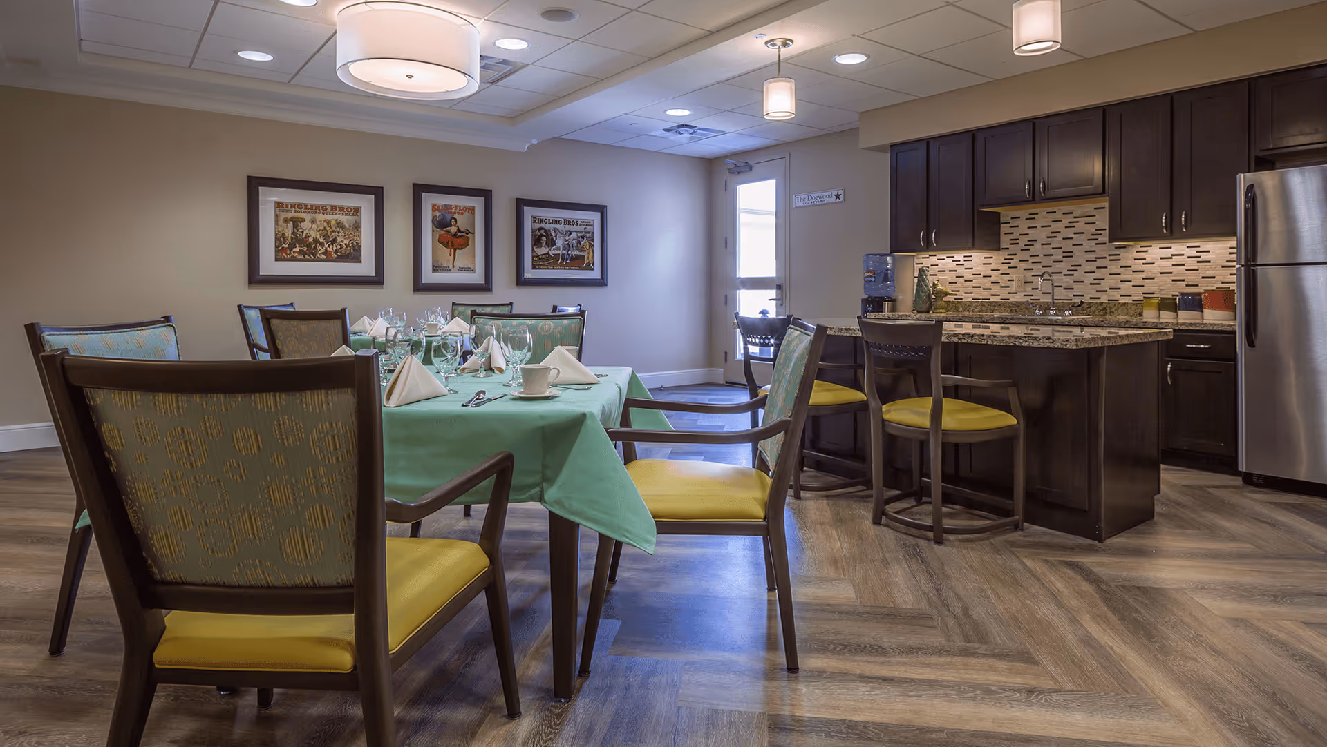 A dining area in a senior living facility with a table covered in a green tablecloth set with napkins, cups, and glasses. Surrounding the table are chairs with yellow cushions and patterned backs. In the background, there is a kitchen area with dark wood cabinets, a granite countertop island with two chairs, a stainless steel refrigerator, and a tiled backsplash. The walls have three framed pictures, and the ceiling features modern light fixtures.