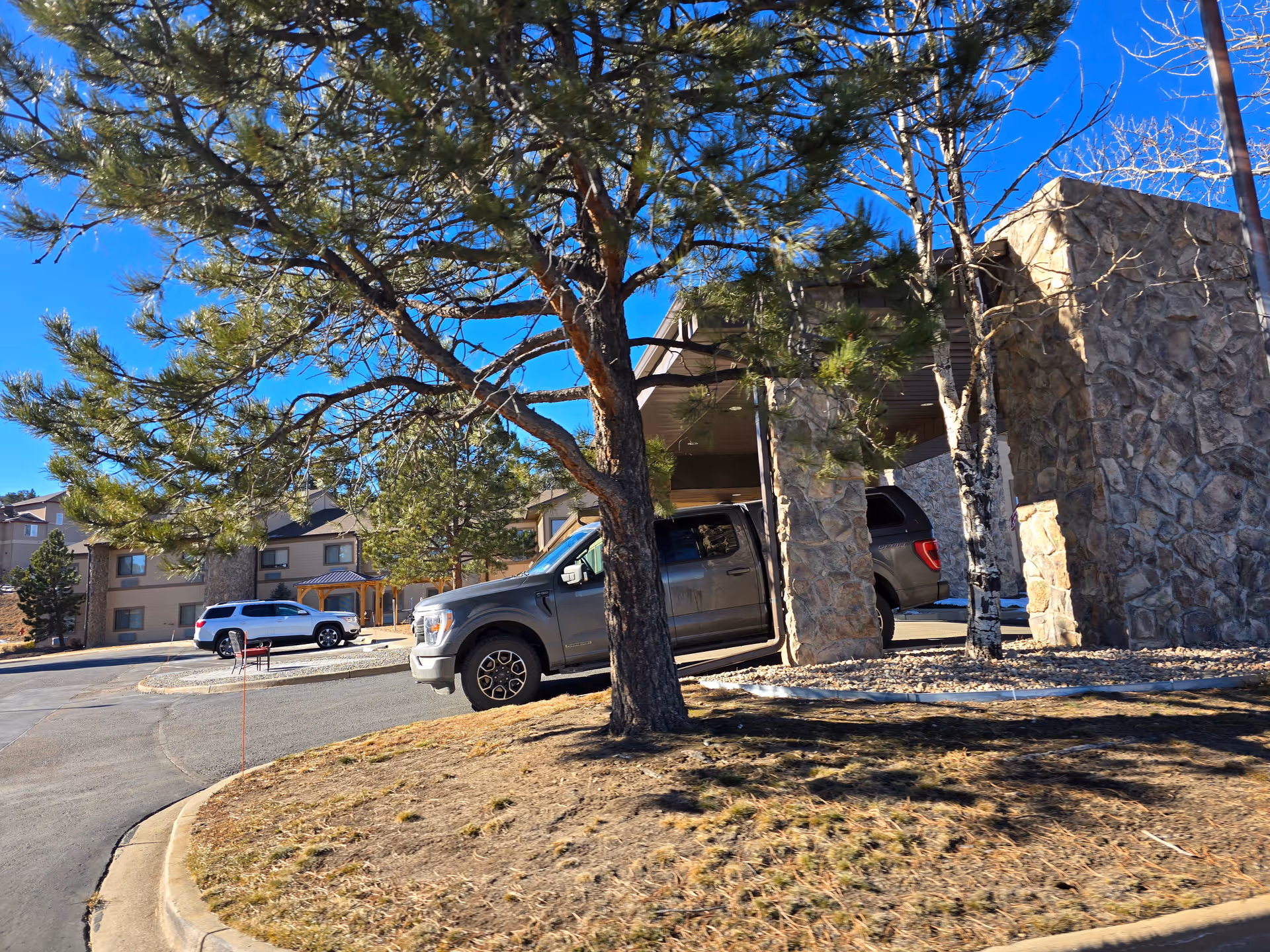 Exterior view of Life Care Center of Evergreen showing a stone entrance canopy with two vehicles parked nearby, surrounded by trees and a clear blue sky.
