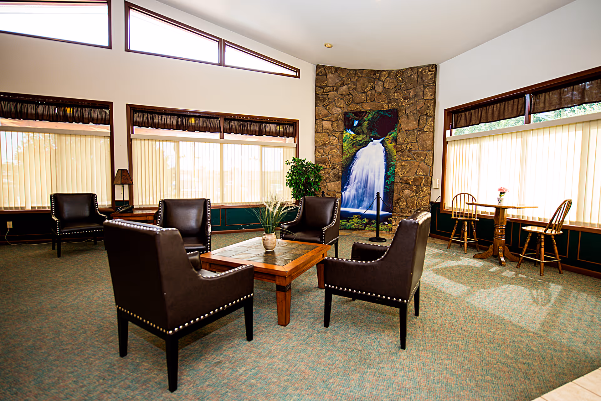 A bright common area with four dark leather chairs arranged around a wooden coffee table with a small plant on it. The room has large windows with vertical blinds, a stone wall with a waterfall painting, and a small round table with two wooden chairs near the window.