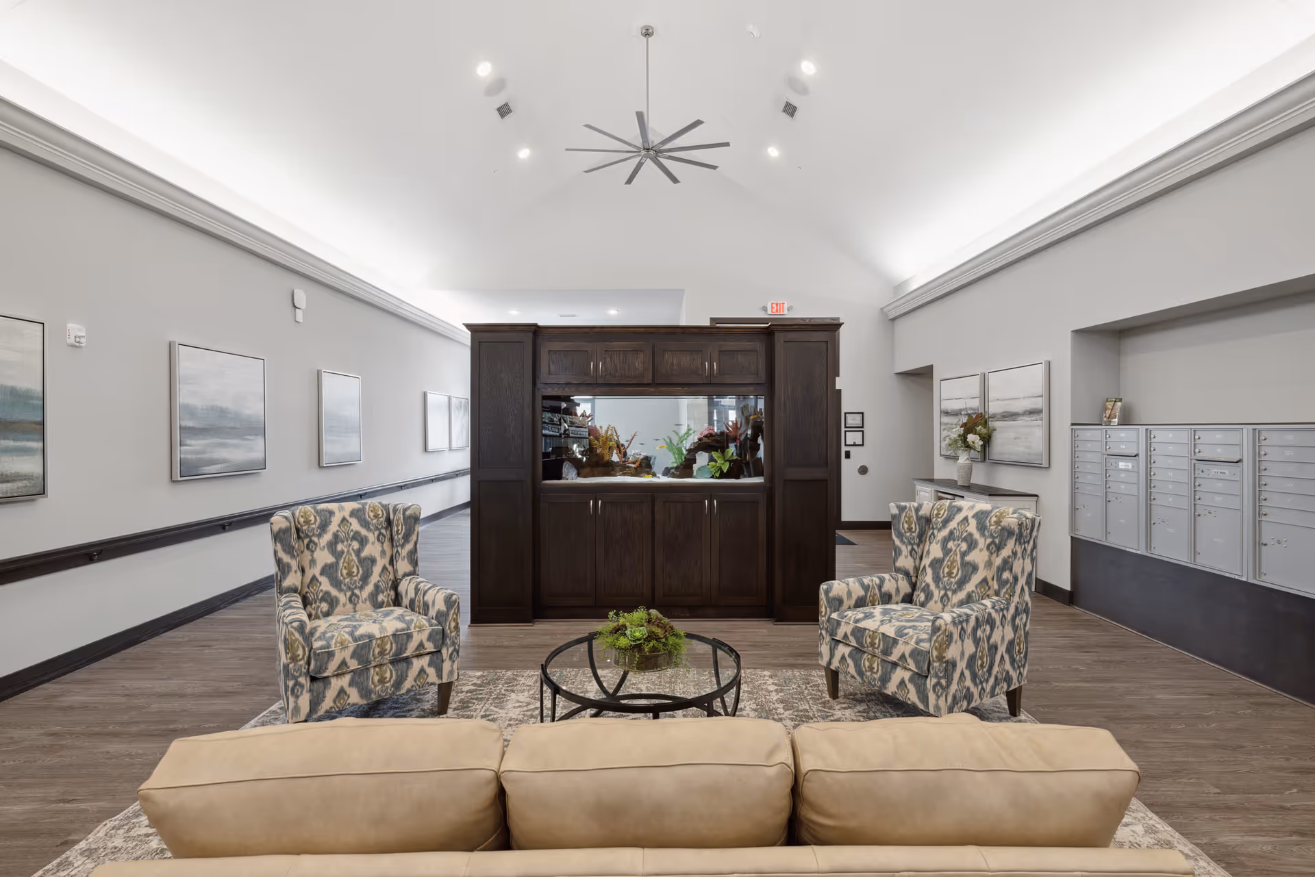 A spacious senior living facility common area with a beige sofa in the foreground, two patterned armchairs facing each other, and a round glass coffee table with a small plant. Behind the seating area is a large dark wood cabinet with an aquarium. The room has high vaulted ceilings with a large ceiling fan, light gray walls adorned with framed artwork, and a row of mailboxes on the right wall.