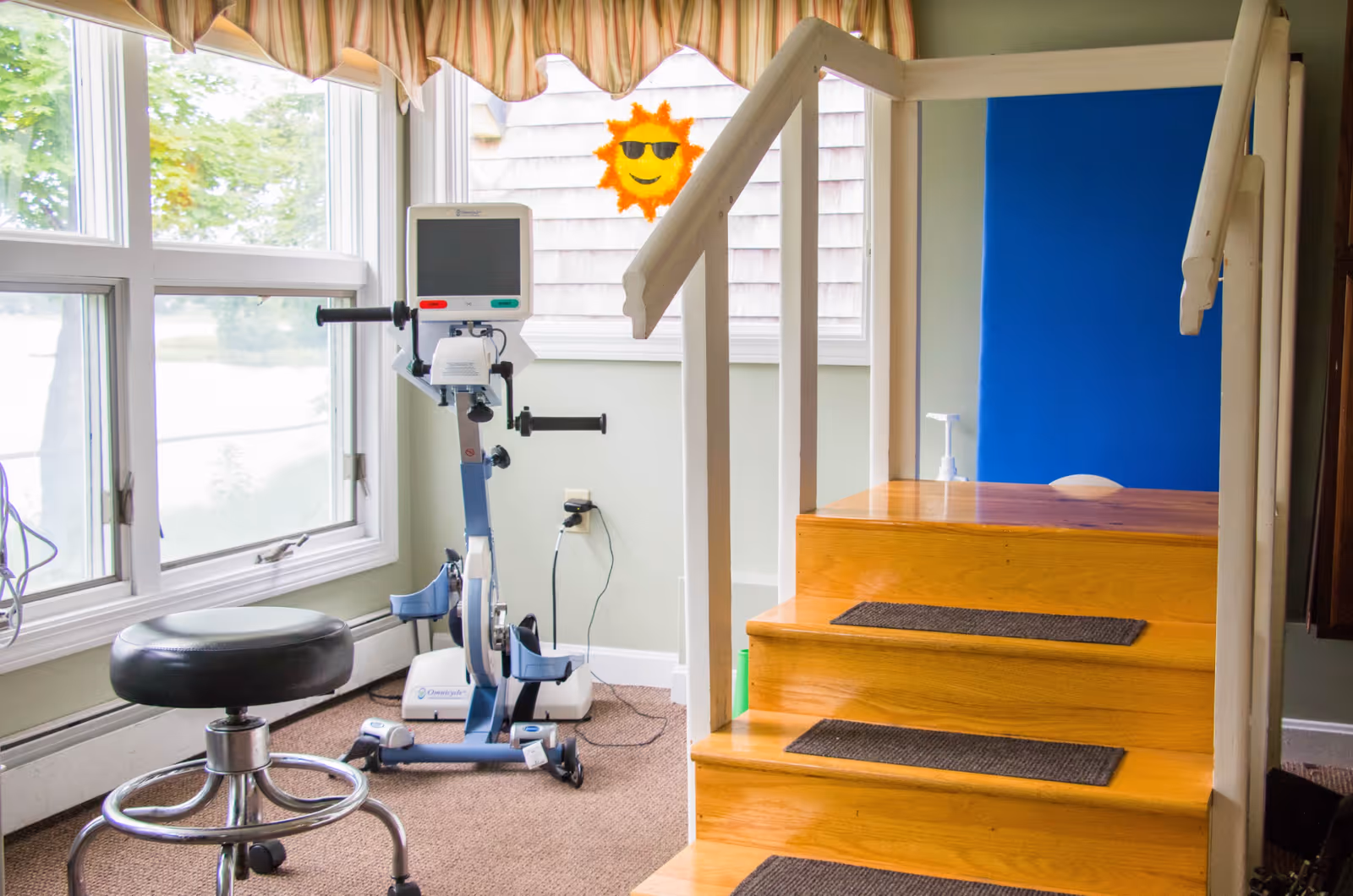 Interior room with a wooden staircase equipped with handrails on both sides, a stationary exercise bike with a digital monitor, a black adjustable stool on wheels, large windows with a striped valance, and a wall decal of a smiling sun wearing sunglasses.