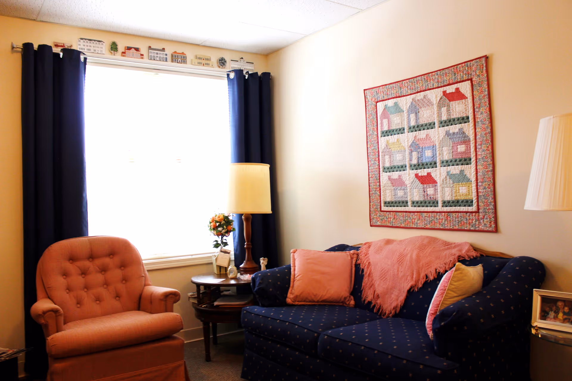 A cozy living room with a blue patterned couch adorned with pink and beige pillows and a pink throw blanket. Next to the couch is a wooden side table with a lamp and a small flower arrangement. There is a coral-colored armchair near a window with dark blue curtains. A colorful quilt with house patterns hangs on the beige wall above the couch.