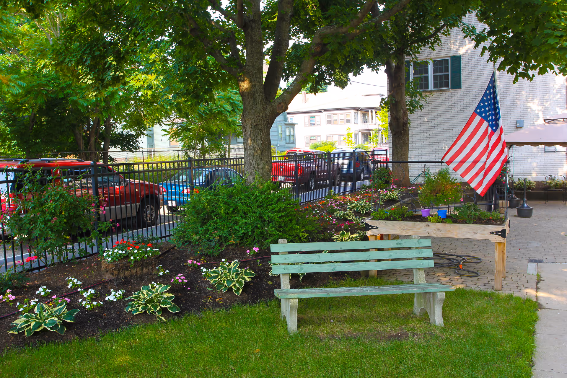 A peaceful outdoor garden area at Elmwood Nursing & Rehabilitation Center featuring a green wooden bench on a grassy lawn, a flower bed with various plants and flowers, a raised garden bed with an American flag, and trees providing shade. In the background, there is a black metal fence and parked cars along the street.