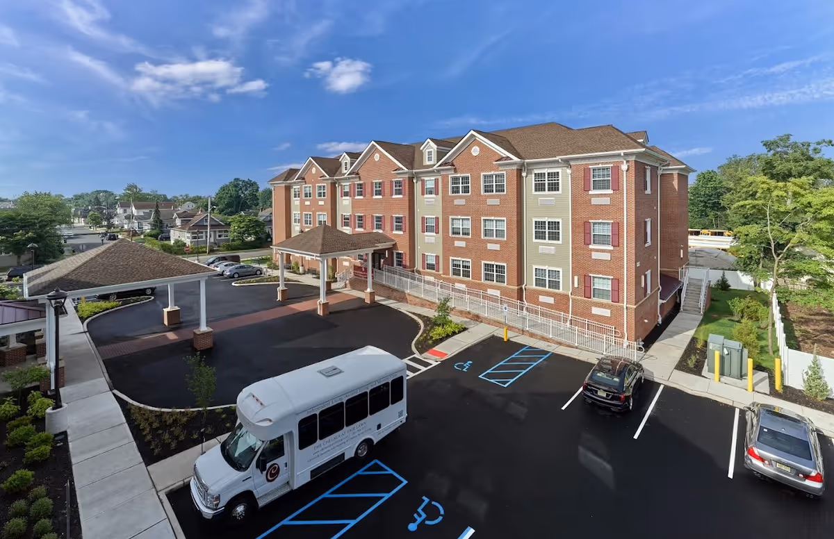 Exterior view of a multi-story senior living facility building with a covered entrance and a parking lot featuring handicap parking spaces and a white shuttle bus. The building has a brick and beige facade with multiple windows and red shutters. The sky is clear with some clouds.