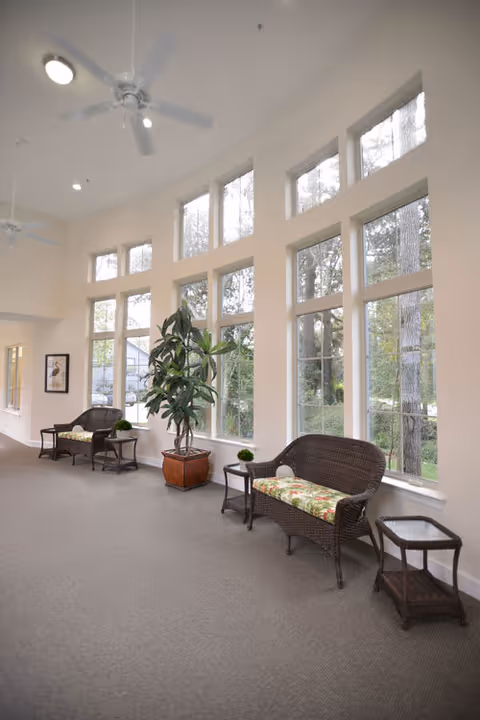 A bright interior lounge area with large windows letting in natural light. The space features two wicker loveseats with floral cushions, small side tables, a large potted plant, and ceiling fans overhead. Outside the windows, trees and greenery are visible.