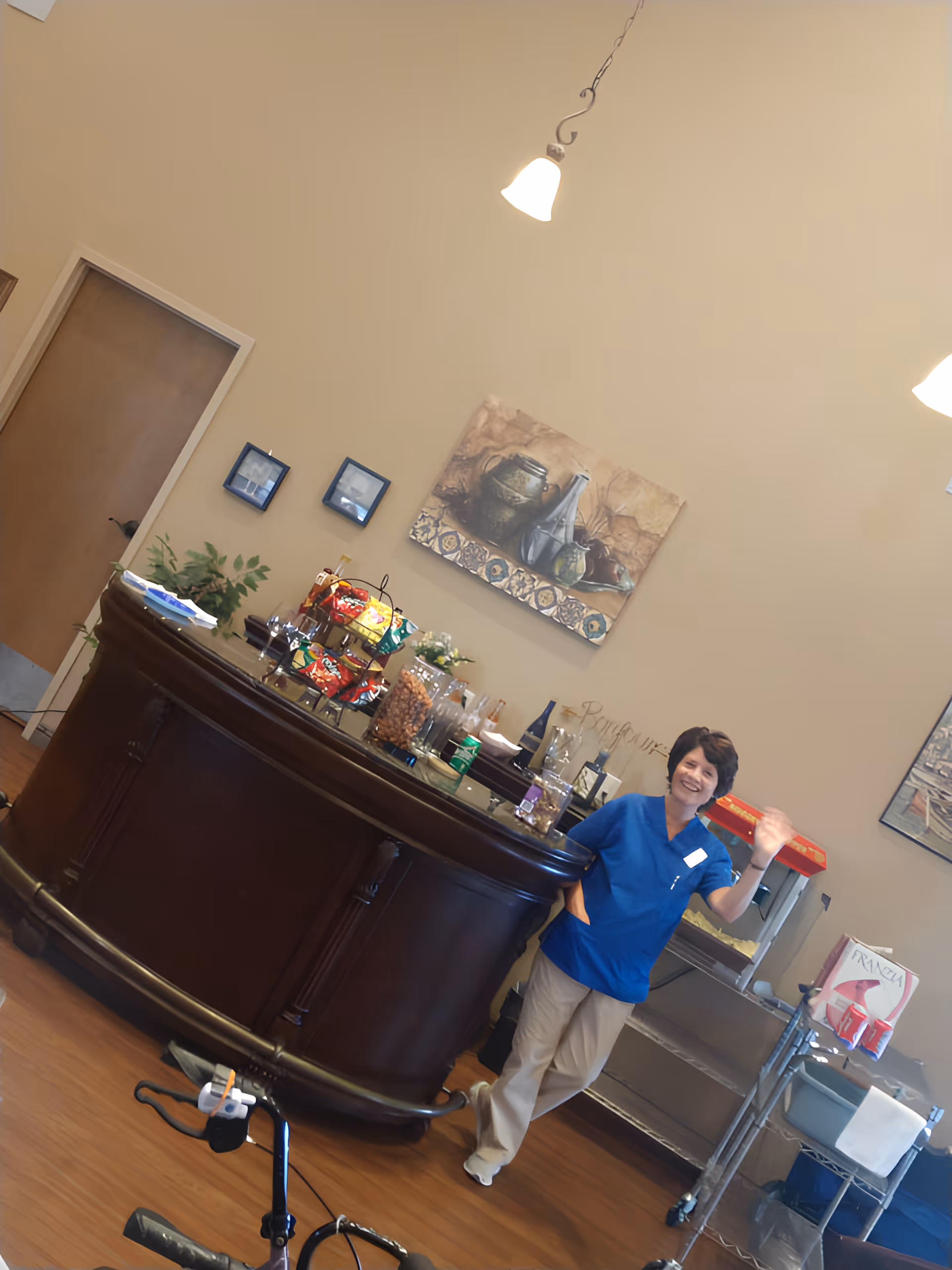 A woman in blue scrubs is standing and waving next to a dark wooden curved counter with snacks and drinks on it. Behind her is a popcorn machine and a metal cart with supplies. The room has beige walls with framed pictures and a hanging light fixture.
