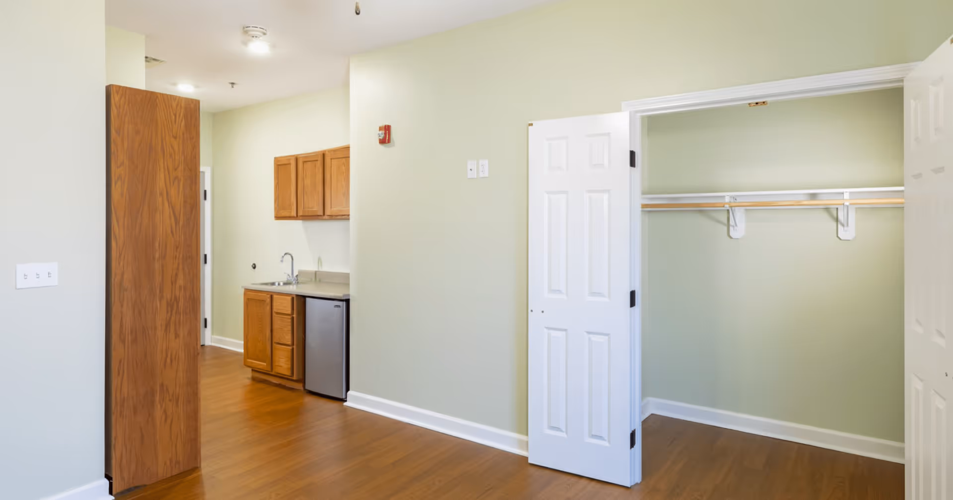 Interior view of a room in an assisted living facility showing a small kitchenette with wooden cabinets, a stainless steel mini refrigerator, and a sink. To the right, there is an open closet with white double doors and a hanging rod. The room has light green walls and wood flooring.