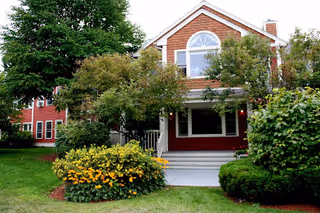 Front exterior of a red two-story house with a porch, surrounded by trees and flowering shrubs.