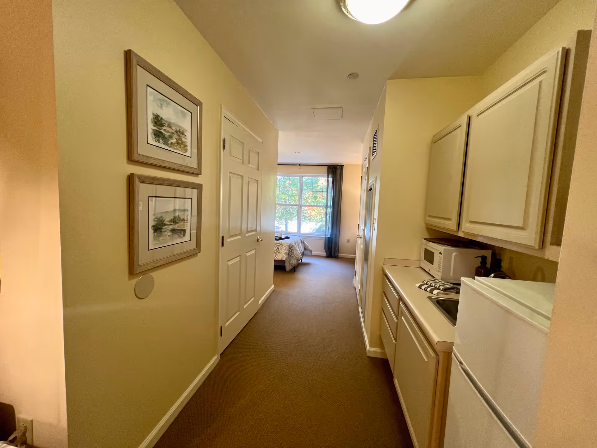 A hallway in a senior living facility with beige walls and carpet. On the right side, there is a small kitchenette area with a microwave, sink, and refrigerator. On the left side, two framed landscape paintings hang on the wall. At the end of the hallway, a bedroom with a bed and a large window with curtains is visible, letting in natural light.