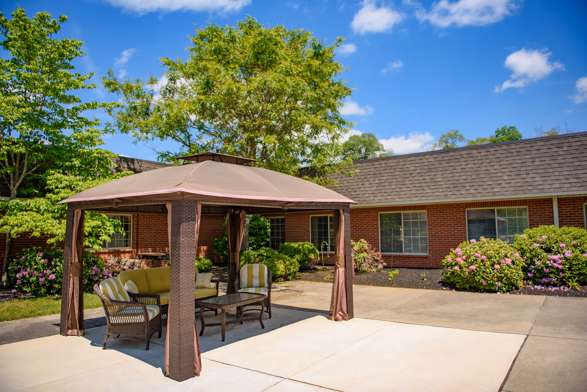 A shaded outdoor seating area with a gazebo and wicker furniture on a paved courtyard in front of a single-story brick building with trees and flowering shrubs.