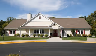 Single-story building with a gable roof and a covered front porch with white railings. The building is surrounded by a well-maintained lawn and shrubs, with a paved driveway and sidewalk leading to the entrance. The sky is partly cloudy.