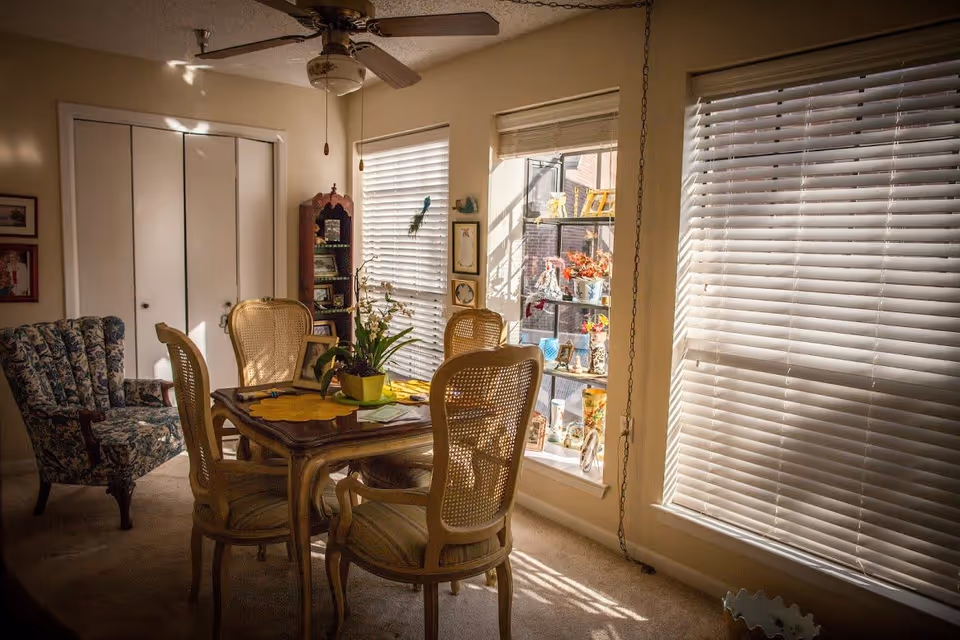 A cozy dining area with a wooden table and four cane-back chairs. The table has a yellow placemat and a potted plant. There is a floral upholstered armchair to the left and a tall wooden corner shelf with framed photos and decorative items. Sunlight streams through two large windows with white blinds, illuminating a display shelf filled with various decorative objects and plants.