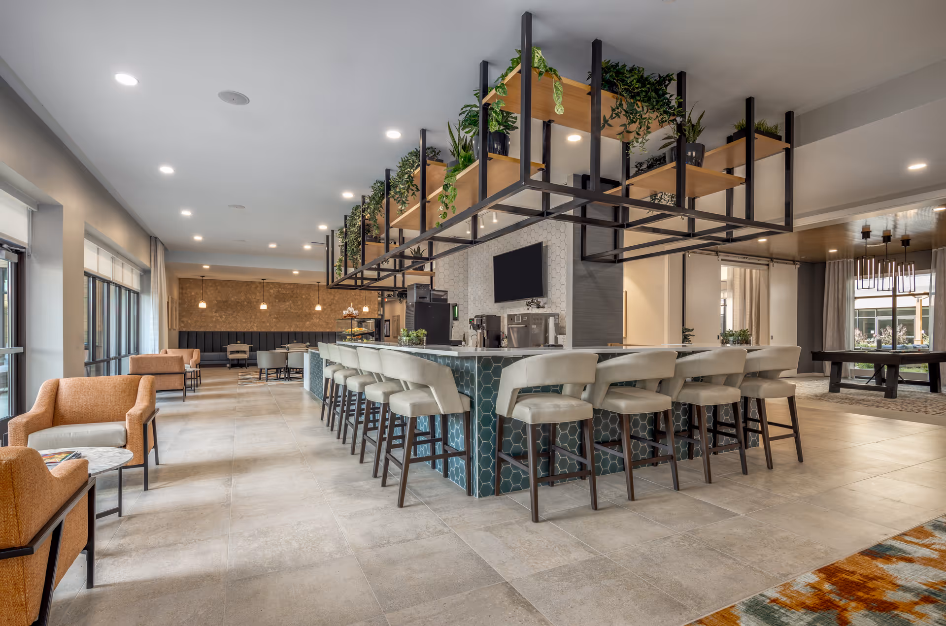 A spacious and modern common area with a large central bar surrounded by cushioned bar stools. Above the bar, there is a hanging shelf with various green plants. To the left, there are comfortable armchairs and small tables near large windows letting in natural light. In the background, there is additional seating and a pool table in a separate section of the room.