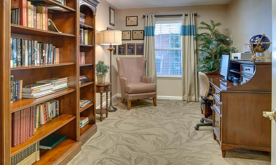 A cozy room with a wooden bookshelf filled with books on the left, a comfortable armchair near a window with curtains, framed certificates on the wall, a floor lamp, a small side table with a plant, and a wooden desk with a computer and a globe on the right.