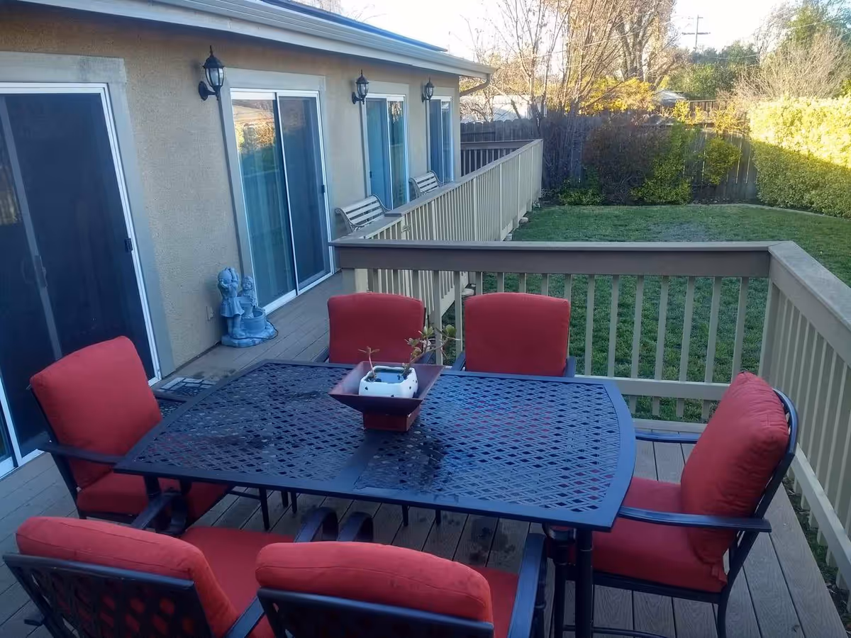 Outdoor patio area with a black metal table and six chairs with red cushions. The patio is adjacent to a building with sliding glass doors and beige walls. There is a small garden area with green grass and bushes enclosed by a wooden fence.