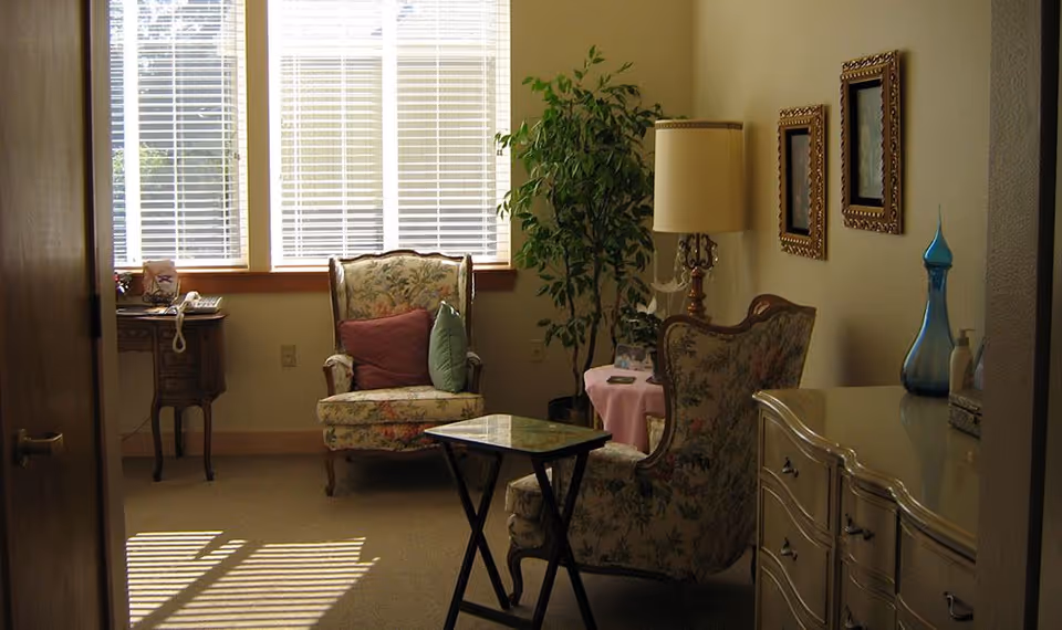 Small sitting room with two floral upholstered armchairs, a lamp and side tables, a dresser with decorative vases, a potted plant, and large windows with blinds.