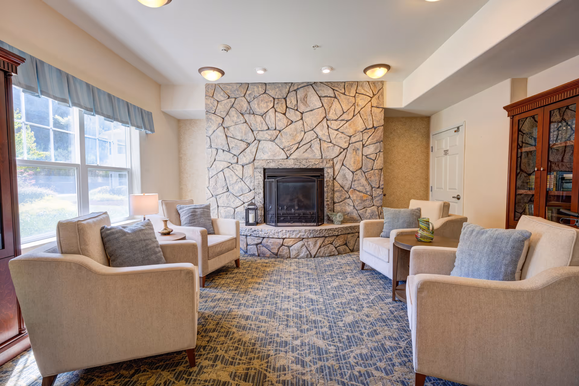 A cozy living room with four beige armchairs arranged around a stone fireplace. The room has a patterned carpet, a large window with blue valance curtains letting in natural light, and a wooden cabinet filled with books on the right side.