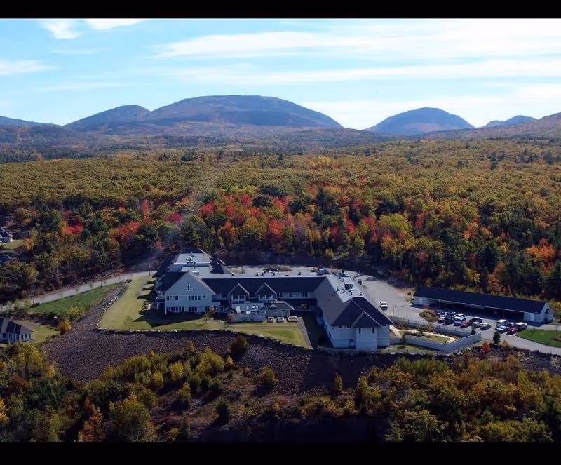 Aerial view of Birch Bay Retirement Village surrounded by dense forest with autumn foliage and mountains in the background under a partly cloudy sky.