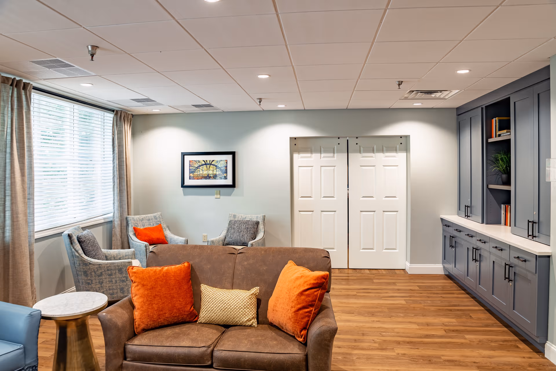 A cozy senior living room with a brown leather loveseat adorned with orange and beige pillows, two gray armchairs with orange pillows, a small round side table, a large window with beige curtains, a framed artwork on the wall, white double doors, and built-in gray cabinetry with shelves holding books and a plant.