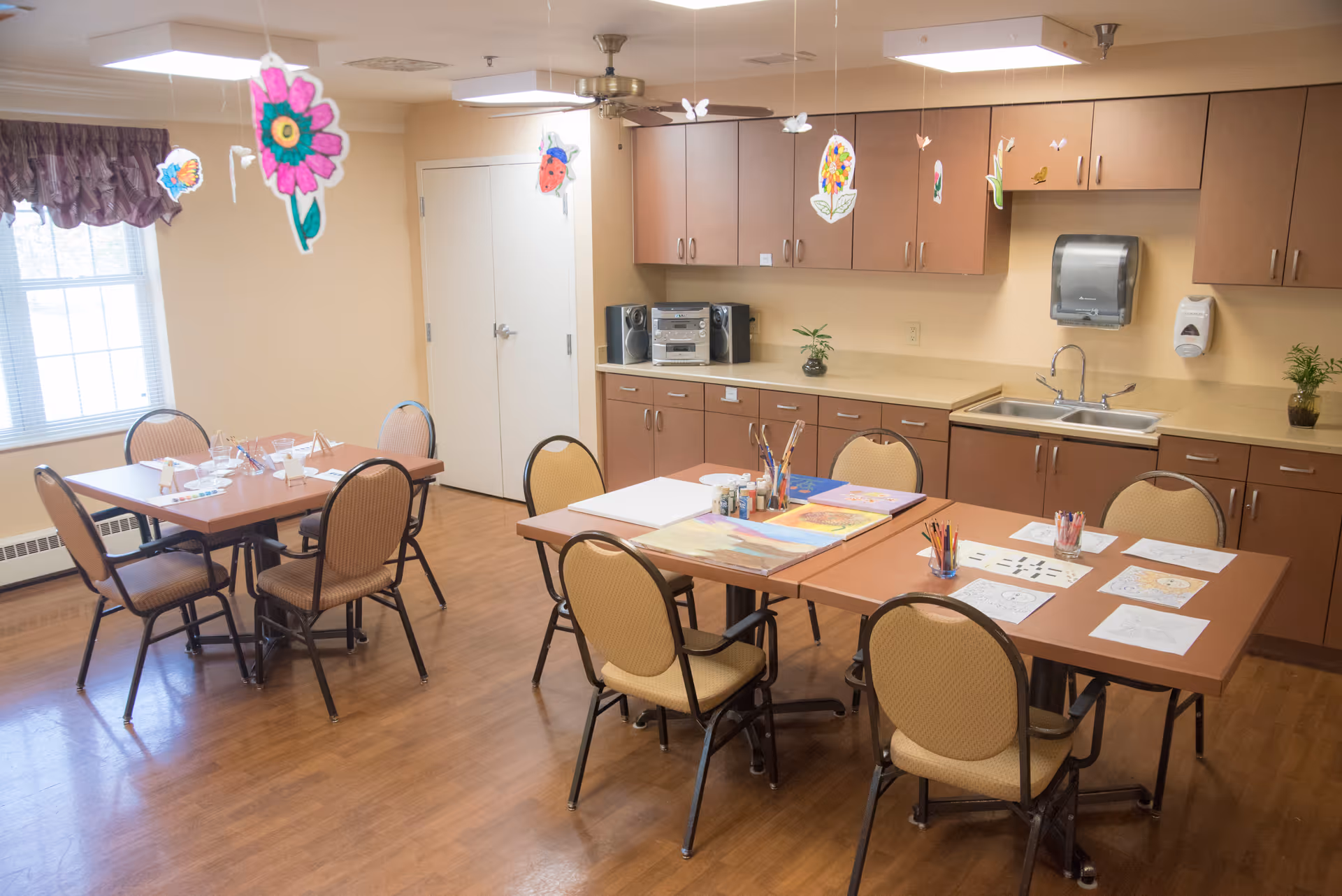 A bright activity room with two tables and several chairs arranged around them. One table has art supplies, coloring pages, and paintbrushes, while the other table is set with glasses and napkins. The room has wooden flooring, beige walls, and a window with a purple valance. Hanging from the ceiling are colorful paper flower and insect decorations. There are brown cabinets along one wall with a sink, a paper towel dispenser, and a stereo system.