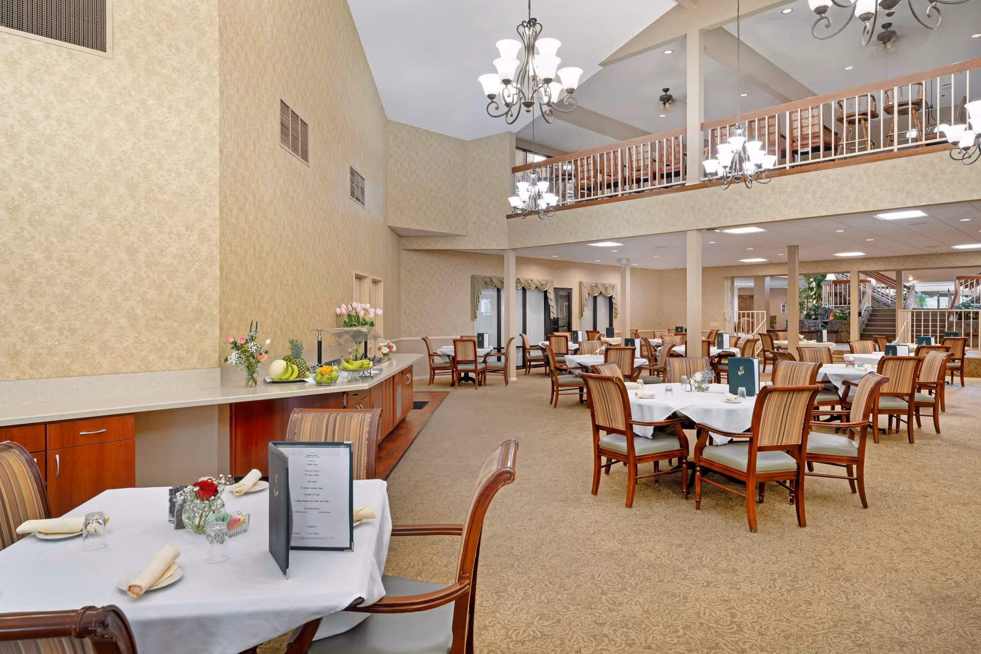 Spacious dining room in a senior living facility with multiple tables covered in white tablecloths, each set with napkins, glasses, and menus. The room features beige patterned wallpaper, carpeted floors, and wooden chairs with striped upholstery. There is a buffet counter with fruit and flowers on the left side, and a mezzanine level with additional seating above. Several chandeliers hang from the ceiling providing warm lighting.