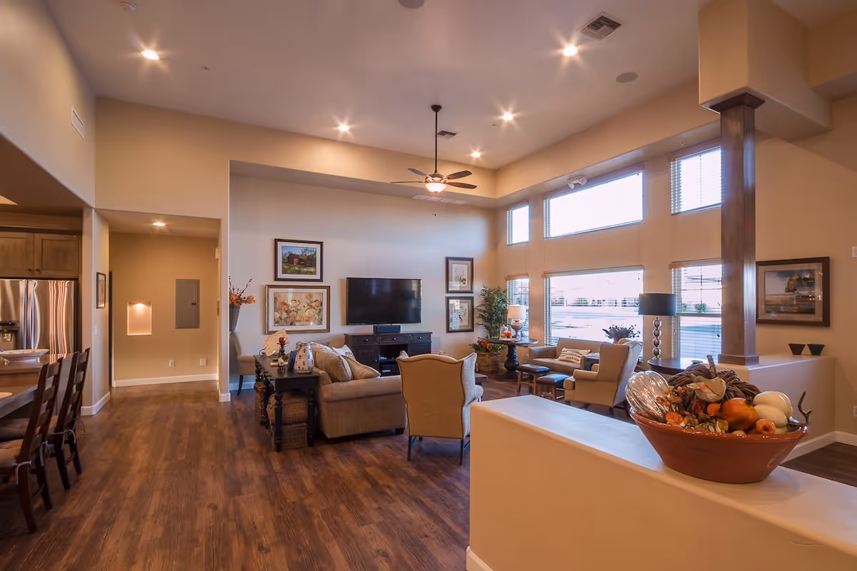 A spacious and well-lit living room area with high ceilings and large windows. The room features comfortable seating including sofas and armchairs arranged around a flat-screen TV mounted on the wall. There are framed artworks on the walls, a ceiling fan, and decorative plants and lamps. The floor is wooden, and part of a kitchen area with a refrigerator and bar stools is visible on the left side.