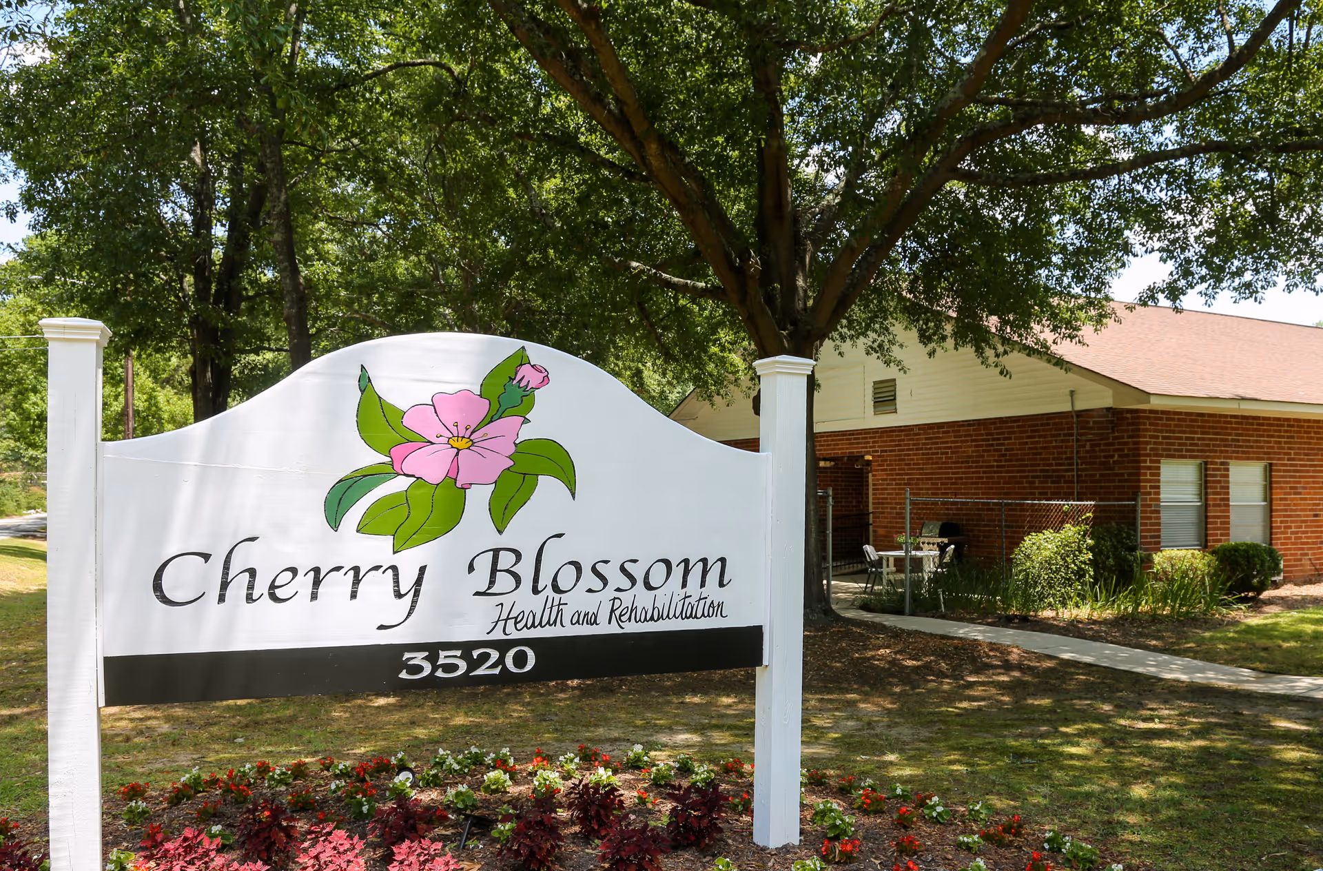 Outdoor view of a white sign with a pink flower illustration that reads 'Cherry Blossom Health and Rehabilitation 3520' in front of a brick building with a tree and landscaped garden.