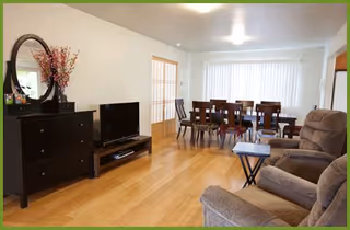 Open living and dining area with hardwood floors, two recliner chairs and a TV in the foreground and a dining table with chairs by a large window.