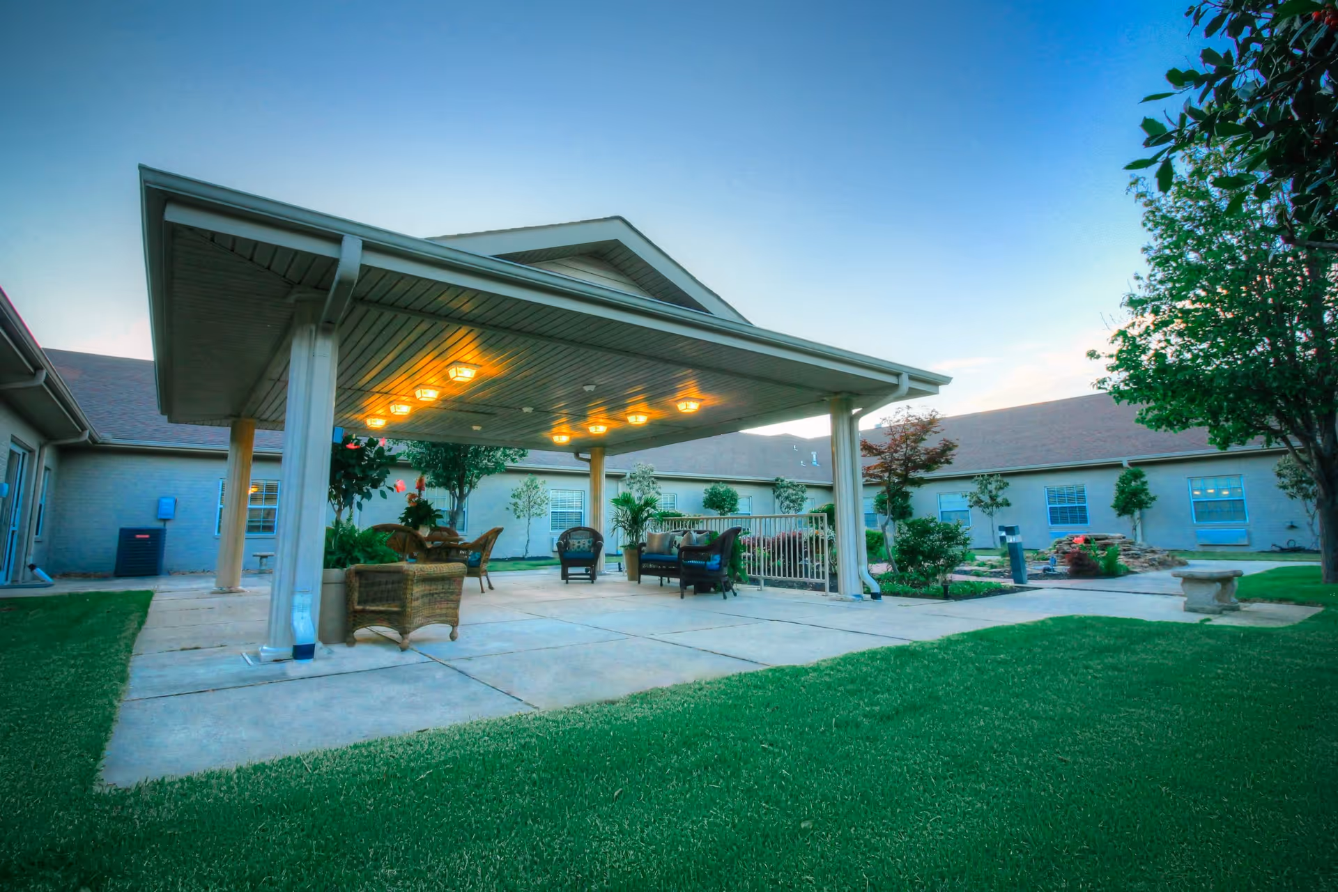 Outdoor covered patio area with seating including wicker chairs and a small table, surrounded by green grass and trees, with a building in the background under a clear sky at dusk.
