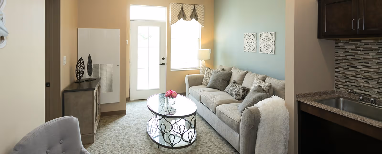 A cozy living room area with a beige sofa adorned with multiple patterned cushions, a glass coffee table with a decorative metal base and a small flower arrangement on top, a side table with a lamp next to the sofa, and a window and glass door letting in natural light. To the right, there is a partial view of a kitchenette with a sink and tiled backsplash. The room has neutral tones and soft carpeting.
