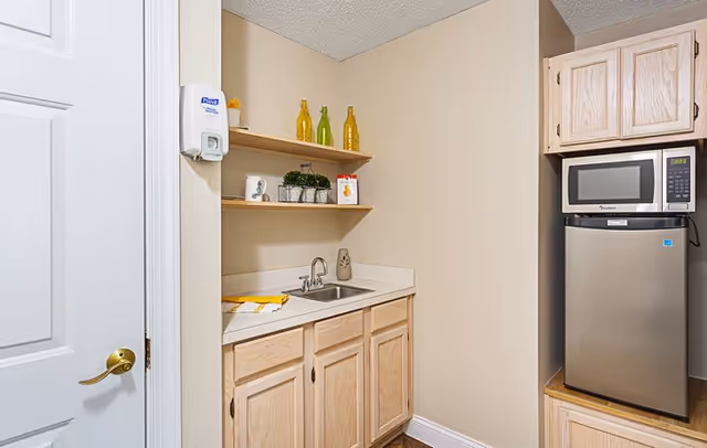 Small kitchenette area with light wood cabinets, a countertop with a sink, two wooden shelves holding decorative bottles and small plants, a microwave on top of a mini refrigerator, and a white door with a gold handle on the left side.