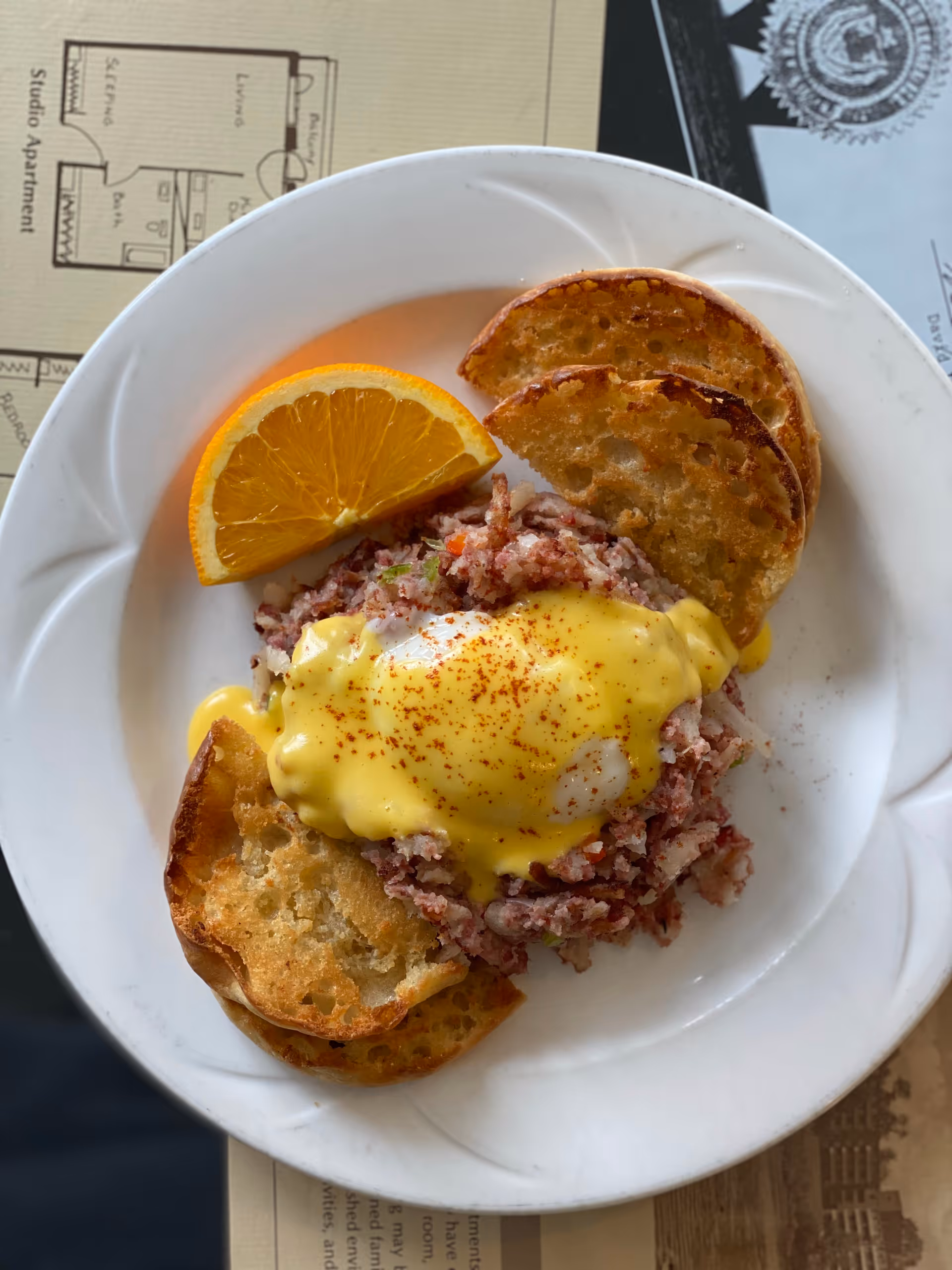 A white plate with a serving of corned beef hash topped with a poached egg and hollandaise sauce, garnished with a sprinkle of paprika. The plate also contains toasted bread slices and a wedge of orange. The plate is placed on top of a paper with a floor plan and some text partially visible underneath.