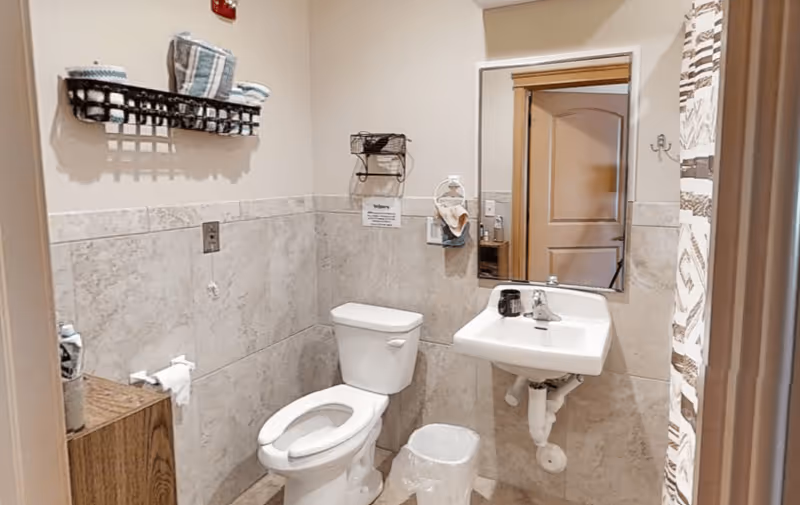 A small bathroom with beige tiled walls and floor, featuring a white toilet, a white wall-mounted sink with a faucet, a mirror above the sink, a small trash can, a towel rack with folded towels, and a shower curtain with a geometric pattern.
