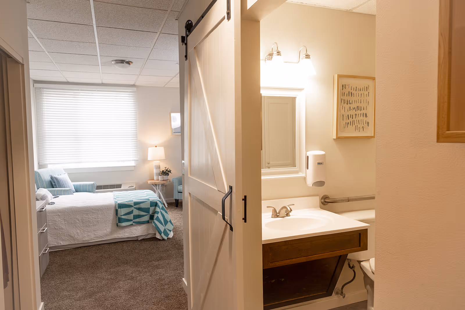 Interior view of a senior living bedroom with a made bed, armchair and bedside lamp, and an adjacent bathroom visible through a sliding barn door.